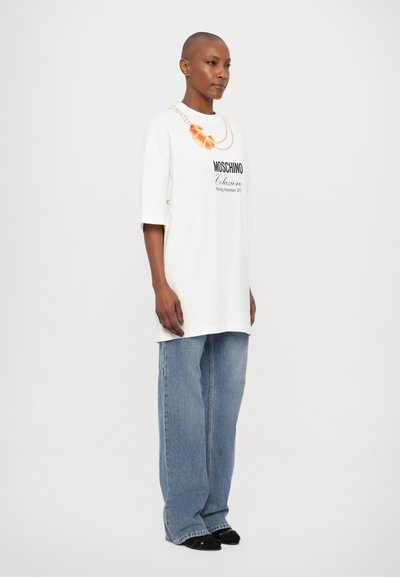 White oversized t-shirt with black Moschino logo print, featuring a decorative necklace graphic. Worn with loose blue denim jeans and black shoes.