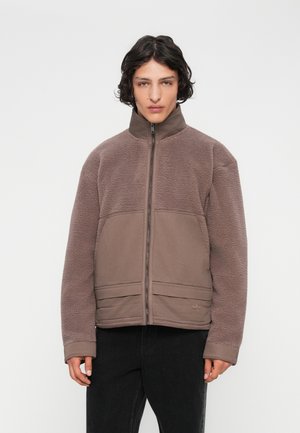 Junge Person mit schulterlangen dunklen Haaren, die eine braune Fleece-Jacke mit Reißverschluss und schwarze Hosen trägt, vor einem weißen Hintergrund stehend.