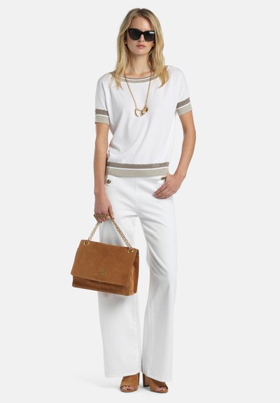 Sieviete baltā topā un biksēs ar bēša apdari, melnās saulesbrillēs, brūnā zamša rokassomiņā ar zelta ķēdi un bēšos atvērtā purngala augstpapēžu kurpēs stāvot.