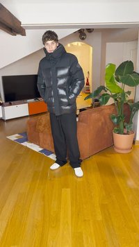 Μαύρο τζάκετ πούφ με γυαλιστερή επιφάνεια και υπερμεγέθη κουκούλα, συνδυασμένο με φαρδιά μαύρα παντελόνια και λευκά αθλητικά παπούτσια, που στέκεται σε ξύλινο δάπεδο.