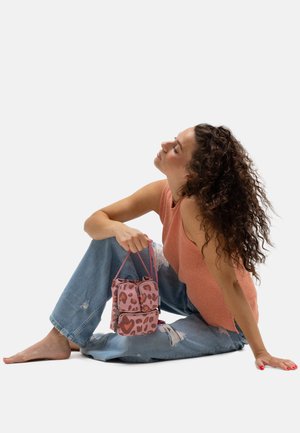 Woman with curly hair sits barefoot on floor, wearing orange sleeveless top and ripped jeans, holding a pink leopard print handbag.