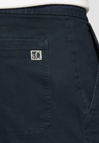 Donker marineblauwe stof met een gladde textuur. Heeft een achterzak en een klein wit "s.O." label op de zak gestikt.