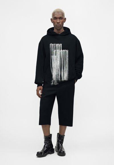 Moški s kratkimi svetlorjavimi lasmi nosi črno kapucar z srebrnim dizajnom cofkov, črne kratke hlače do kolen in sijoče črne gležnarje.