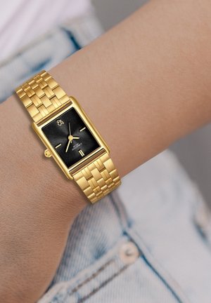 Rechteckige goldene Uhr mit schwarzem strukturiertem Zifferblatt und goldenen Stundenmarkierungen. Goldener geflochtener Armband. Am Handgelenk eines hellen Hauttons getragen.