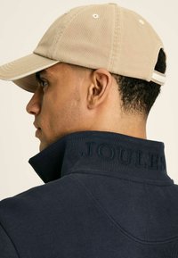 Jūras zila krekls ar paceltu "JOULES" izšuvumu pie kakla, apvienots ar bēša cepuri ar izliekto malu un regulējamu siksnu.