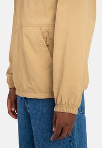 Lys beige letvægtsjakke med elastiske manchetter, en lynlåslomme i siden og en afslappet pasform, parret med blå denim jeans.