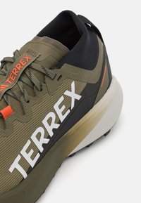 Terrex batai turi žalią audinį viršutinę dalį su baltais logotipais, juodais akcentais ir tekstūruota padanga. Dizainas apima išreikštą paminkštintą kulną.
