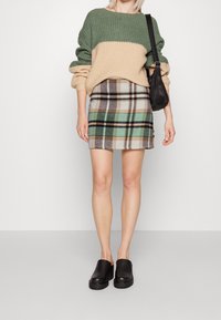 Chunky groene en beige gebreide sweater met brede mouwen, gecombineerd met een geruite minirok in tinten beige, groen en zwart. Zwarte instapschoenen.