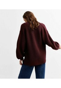 Burgundy stickad tröja med lös passform, ribbat mönster och ballongärmar. Tillsammans med blå denimjeans, visande bakifrån.