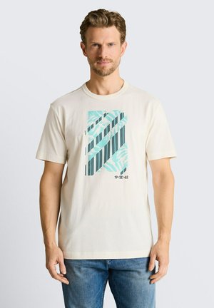 Man met lichtbruin haar die een crèmekleurig T-shirt draagt met een blauw en zwart gestreept abstract ontwerp en een blauwe spijkerbroek, staand tegen een witte achtergrond.
