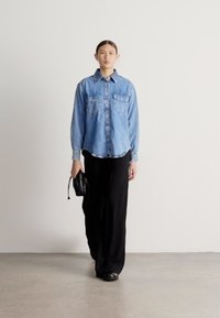 Svetlomodrá rifľová košeľa s dvoma náprsými vreckami, kombinovaná s čiernymi širokými nohavicami a malou čiernou kabelkou. Jednoduchý, minimalistický dizajn.