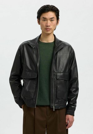 Veste en cuir noir avec fermeture éclair devant, deux poches poitrine et poignets côtelés. Portée par-dessus un pull vert, présentée sur un fond uni.