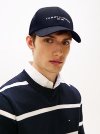 Tengeri kék baseball sapka, rajta fehér hímzéssel "TOMMY HILFIGER EST. 1985" felirattal. Kék pulóverrel viselve, amelyen fehér vízszintes csíkok találhatók.