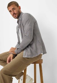 Camicia con bottoni a quadri blu e marrone, abbinata a pantaloni beige e sneakers bianche. Sgabello in legno naturale.
