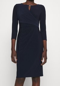 Mörkblå knälång klänning med trekvartsärmar, asymmetrisk draperad kavaj och en liten spänndetalj vid halsringningen.