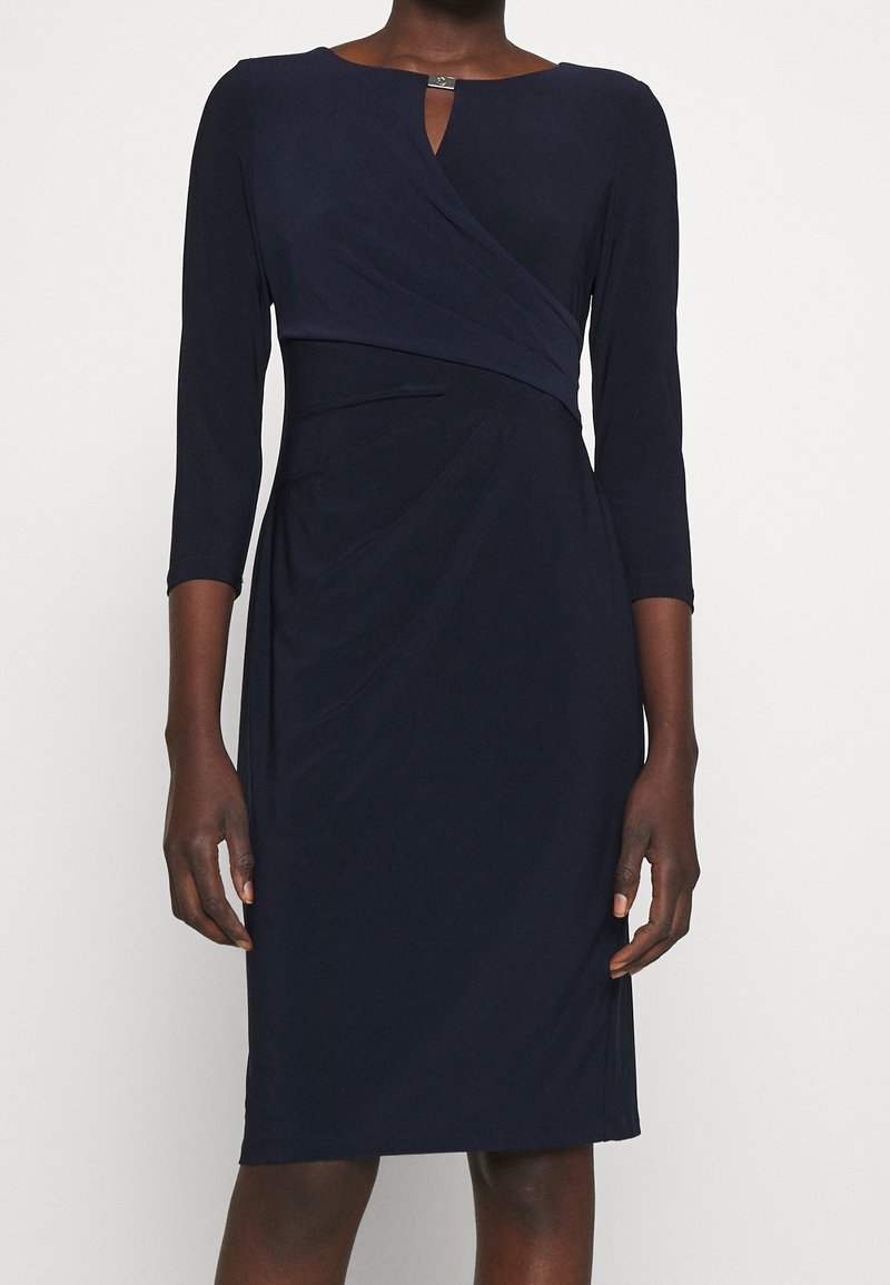 Mörkblå knälång klänning med trekvartsärmar, asymmetrisk draperad kavaj och en liten spänndetalj vid halsringningen.