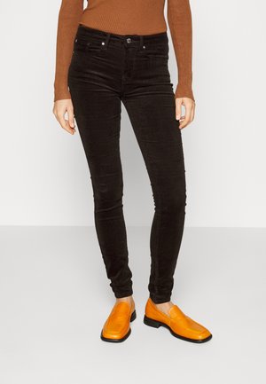 Frau trägt schwarze Cordhosen in Slim-Fit, braunes geripptes Langarmshirt und leuchtend orangefarbene Lederloafer, steht auf hellem grauem Boden.