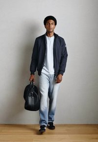 Sort bomberjakke, hvid t-shirt, lyseblå jeans og sorte sneakers. Bærer en sort træningstaske. Glatte teksturer og afslappet design.
