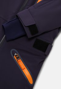 Granatowa kurtka softshell z kontrastowym pomarańczowym zamkiem błyskawicznym i detalami w postaci pasków. Posiada mankiety na rzepy oraz gładką teksturę. Zawiera funkcjonalne elementy projektowe.