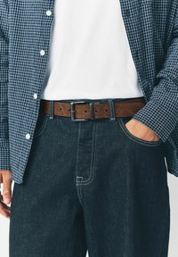 Cinturón de cuero marrón con hebilla de metal, usado con pantalones de mezclilla oscuros y una camisa a cuadros azul sobre una camiseta blanca.