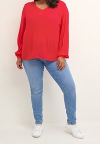 Blouse rouge à manches longues avec un col en V et une texture douce et fluide. Associée à un jean bleu clair et des baskets blanches.