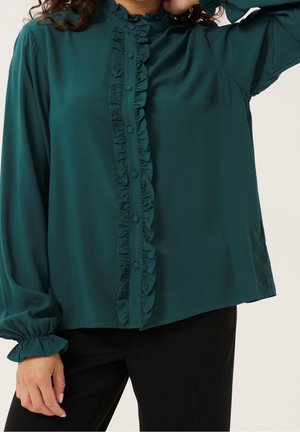 Mujer que lleva una blusa de manga larga de color azul oscuro con volantes y botones cubiertos de tela en la parte delantera, combinada con pantalones negros.
