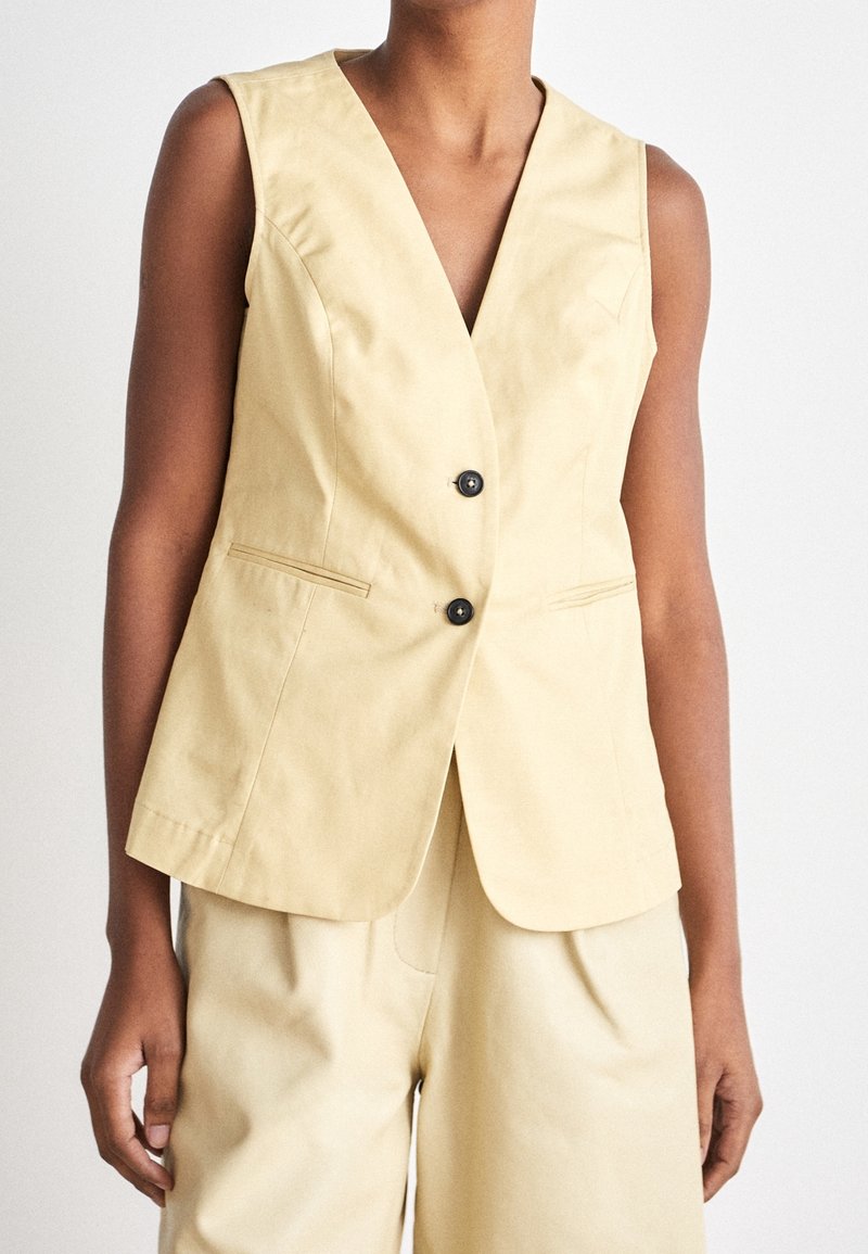 Blazer beige sans manches avec deux boutons noirs et short taille haute assorti porté par une personne à la peau de teint moyen-foncé.