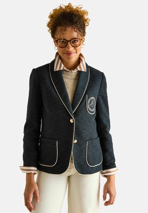 Dunkelgrauer Tweed-Blazer mit weißer Paspel, zwei Vordertaschen und goldenen Knöpfen. Darunter ist ein cremefarbener Pullover mit gestreiftem Kragen sichtbar.