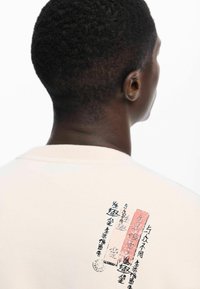 Krém színű pamut pulóver, fekete és piros színű szöveges és grafikus nyomattal a hátsó oldalon, kerek nyakkivágással és puha tapintással.