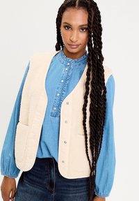 Gilet en polaire beige avec des boutons pressions et deux poches avant, porté par-dessus une chemise en denim bleu avec un col volé et des manches longues.
