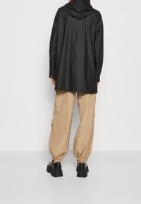 Czarna wodoodporna kurtka z kapturem, o opływowym kroju i zapięciu na zatrzaski, w zestawieniu z luźnymi beżowymi spodniami i masywnymi czarnymi butami.