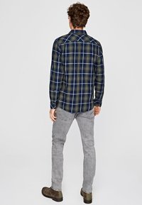 Camicia flanella a quadri verde scuro e blu navy, con colletto a bottoni e maniche lunghe. Indossata con jeans grigi e stivali marroni.