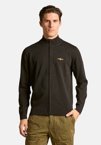 Maglione nero con zip frontale e collo alto, con logo dorato sul petto. Realizzato in tessuto knit liscio con polsini e orlo a costine.