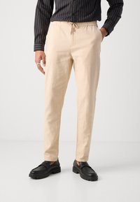 Beige leichte Hose mit strukturierter Oberfläche, elastischem Bund mit Kordelzug und eng zulaufendem Schnitt. Kombiniert mit schwarzen Slippers.