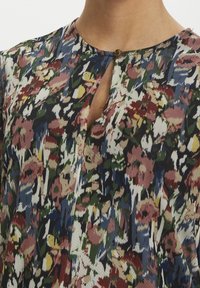 Bluse mit floralem Muster, rundem Ausschnitt, kleinem Schlüsselhalsteil und braunem Knopf-Detail. Fertiggestellt in lebendigen Farben wie Blau, Pink und Grün.