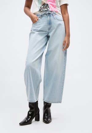 Person trägt hellblaue Jeans mit weitem Bein, schwarze knöchelhohe Stiefel mit Absatz und ein bunt gemustertes T-Shirt, steht mit einer Hand in der Tasche.