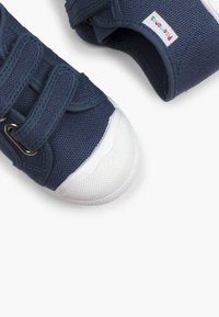 Tumedat sinised kangast jalatsid valgete kummist varbakatetega ja tekstuursete tallad, millel on kolm Velcro pandlat ja värviline brändimärk.