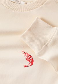 Sweat-shirt couleur crème avec poignets côtelés et petit motif crevette rouge près du poignet sur tissu doux.