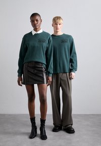 Two models stand side by side. The first wears a dark green ribbed sweater with a logo, a white collared shirt, and a black leather mini skirt. The second model wears a matching green sweater and loose gray trousers. Both have black footwear.