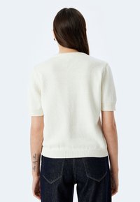 Maglione a maniche corte in maglia di colore avorio, con scollo e orlo a costine. Indossato con jeans scuri, presenta una texture semplice e un design pulito.