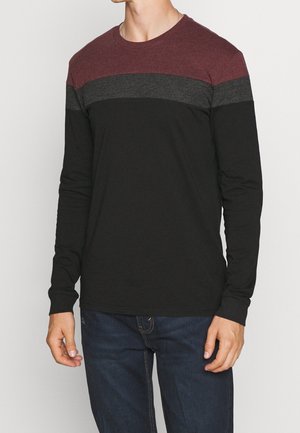 Mężczyzna ubrany w koszulę z długim rękawem z poziomymi blokami kolorów w bordowym, ciemnoszarym i czarnym, w połączeniu z ciemnoniebieskimi dżinsami.