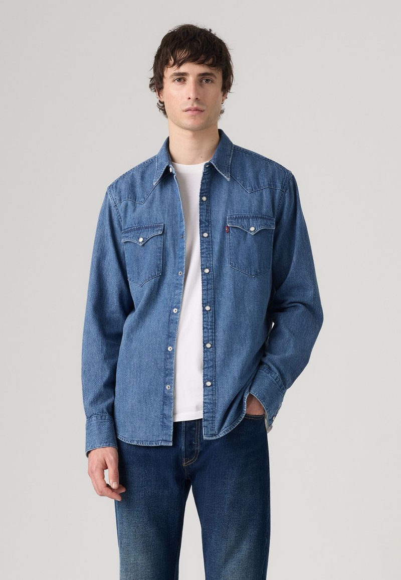 Jeune homme portant une chemise en jean bleue à boutons sur un t-shirt blanc et un jean bleu, debout devant un fond clair uni.