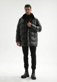 Zwarte pufferjas met een glanzende afwerking, grote doorgestikte delen en een met bont gevoerde kraag, gedragen over donkere kleding en zwarte laarzen.