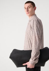 Borsa in pelle nera tenuta da una persona che indossa una camicia rosa chiaro con motivi neri e bianchi sparsi. La borsa ha una forma arrotondata e morbida.