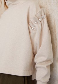 Béžový cropped svetr s vysokým límcem, s nařasenými rukávy zdobenými šesti velkými umělými perlami. Měkká látka s hladkou texturou.