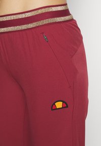 Vinröda träningsbyxor med elastisk midja i guldstrimmor, som har en sidoficka med dragkedja och en cirkulär logotyp med röda, gula och svarta inslag.
