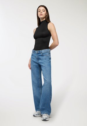 Bodycon senza maniche nero, jeans a gamba larga azzurri con vita alta, sneakers argentati e texture lisce su uno sfondo neutro.