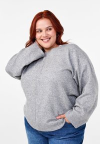 Svetlo sivi oversized pulover z okroglo izrezom, rebrastimi manžetami in gladko teksturo. Nosi se z modrimi kavbojkami, kar poudarja sproščen videz.