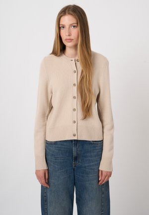 Young woman with long straight hair wearing a beige buttoned cardigan and blue jeans, standing against a plain light background.