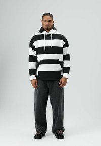 Sweat à capuche à rayures noires et blanches avec cordon de serrage, poche avant et poignets côtelés, associé à un jean large foncé et des bottes noires à lacets.